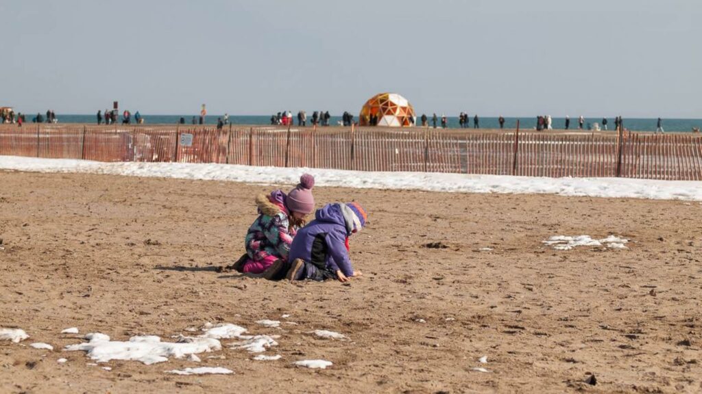 Kids are playing with sand at the Beaches in Toronto. Winter Stations installation and visitors are the background. 